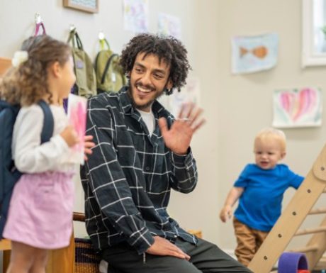A parent kneels to hug a toddler goodbye while a smiling teacher waits nearby in a bright Willowdale classroom